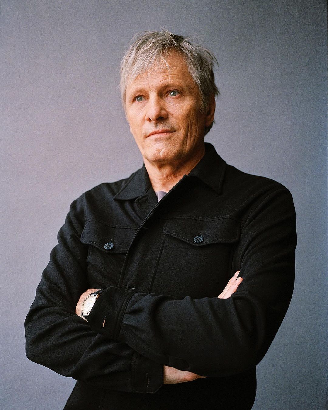 Portrait of an adult man with gray hair and a black button-down shirt, standing with his arms crossed against a gray background.