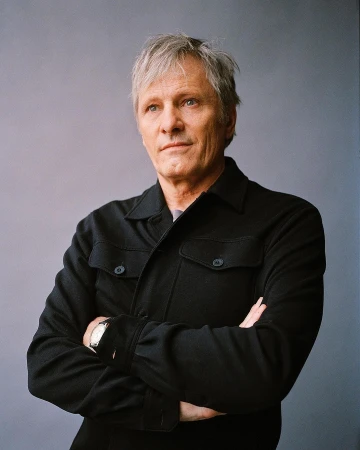 Portrait of an adult man with gray hair and a black button-down shirt, standing with his arms crossed against a gray background.