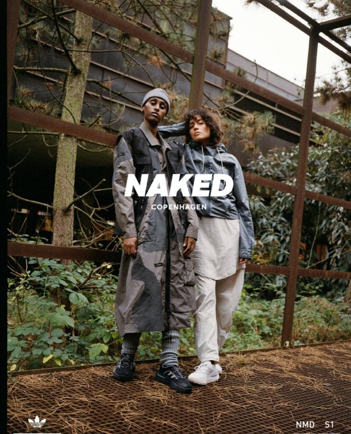 Two women in a fashion ad wearing coordinated outerwear and sneakers. Shot outdoors on a rusted metal structure with green foliage background.