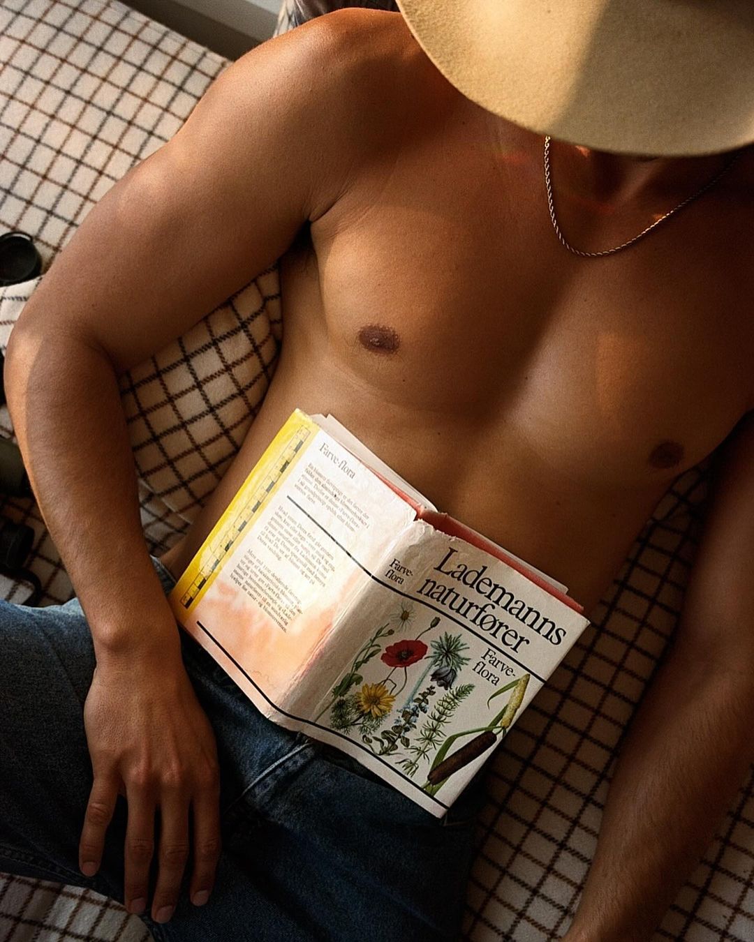 Adult male torso and legs in jeans reclined on bed, reading a book; wearing a hat, natural light. Wellness and literature concept.