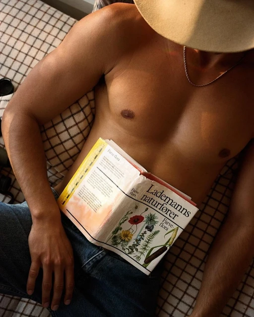 Adult male torso and legs in jeans reclined on bed, reading a book; wearing a hat, natural light. Wellness and literature concept.