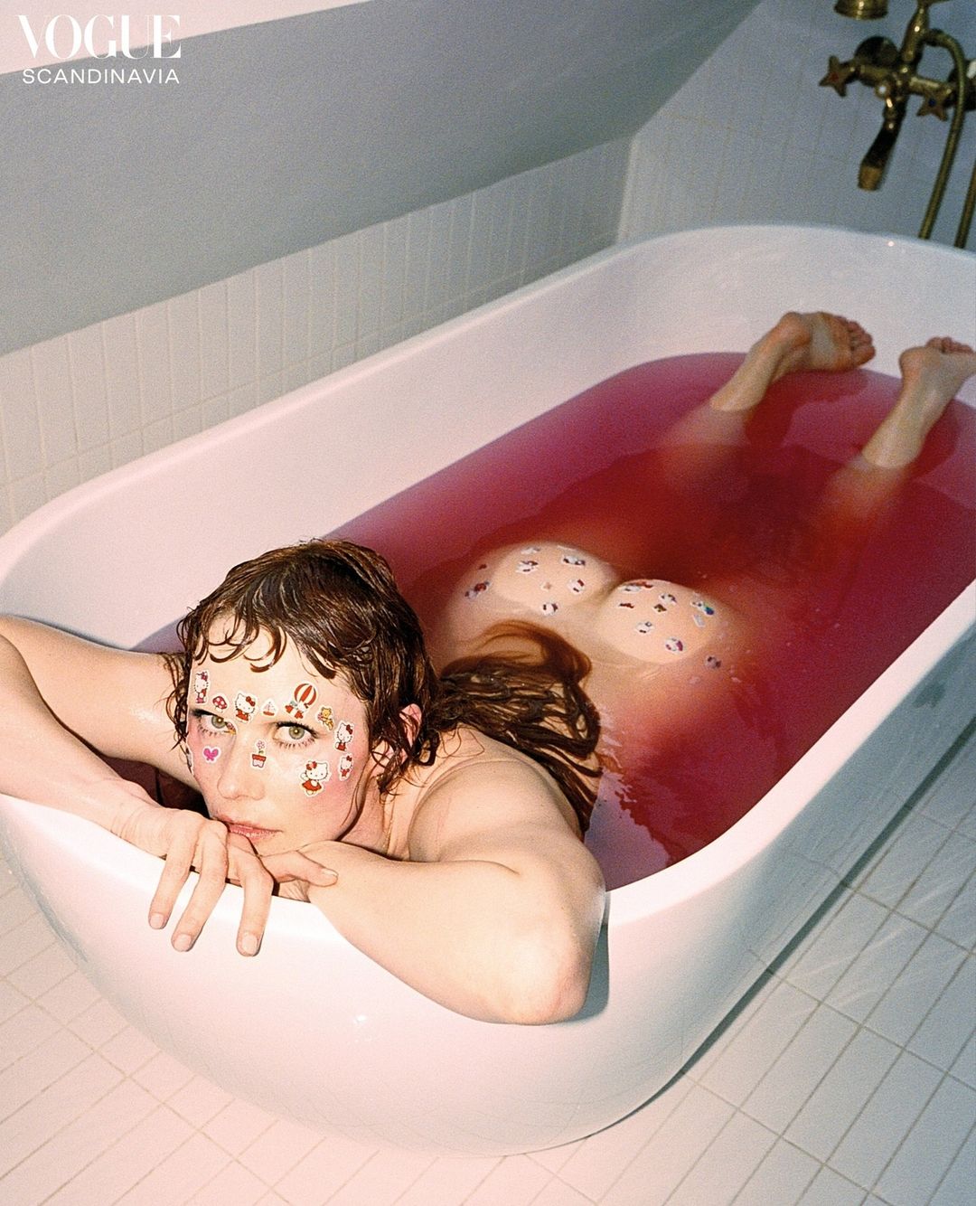 Woman in bathtub with red water and stickers on her face, leaning her head on the edge of the tub. Vogue Scandinavia logo in corner.