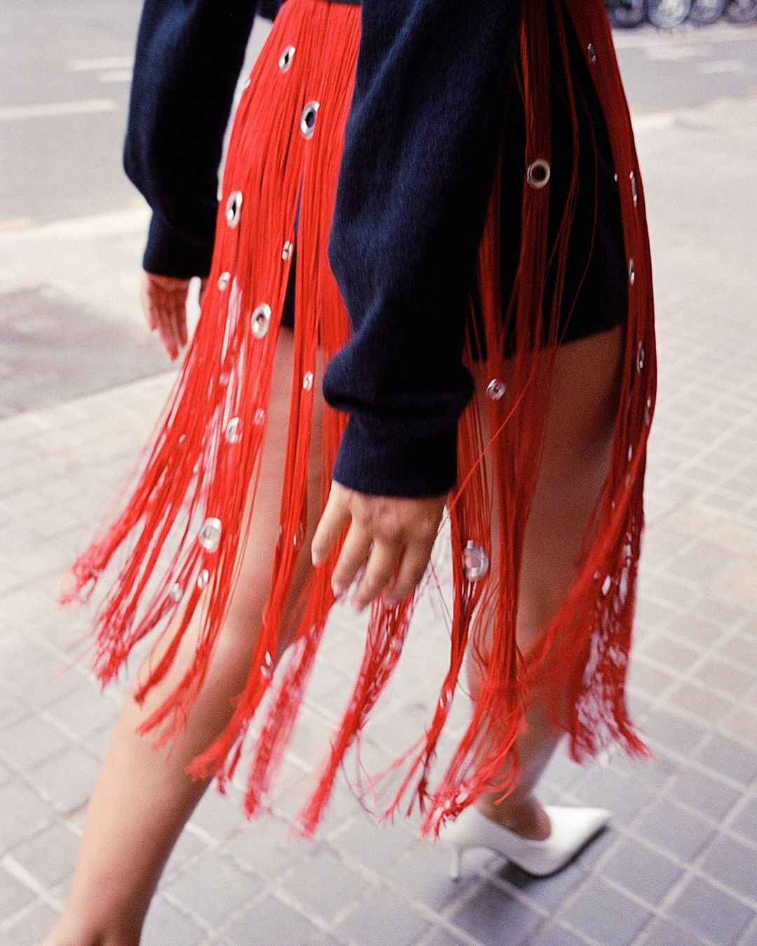 A woman wears a fringed skirt with silver grommets paired with white heels in an outdoor setting, showcasing street fashion.