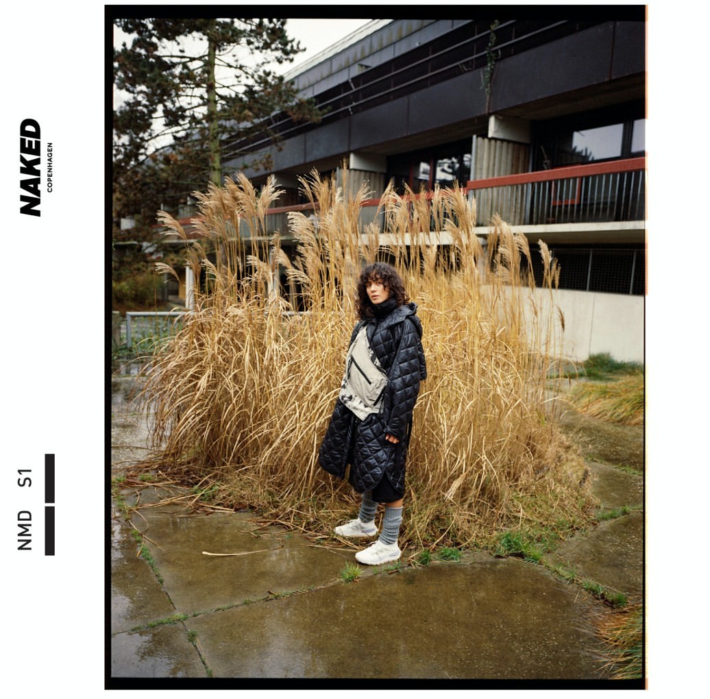 A fashionable woman wearing a long black puffer coat and a beige bag stands in front of tall reeds, with a building in the background.