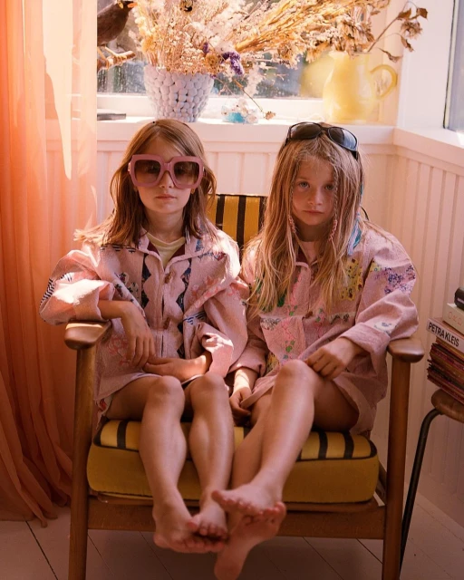 Two young girls wearing matching jackets and sunglasses sit closely together in a chair indoors bathed in warm light.
