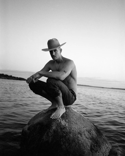 Black and white portrait of a man with a hat, bare-chested, sitting on a rock in the water. Ocean background with a horizon line.
