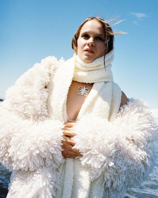 Woman in a fluffy white robe and scarf stands against a blue sky with water in the background, posing with hands together.