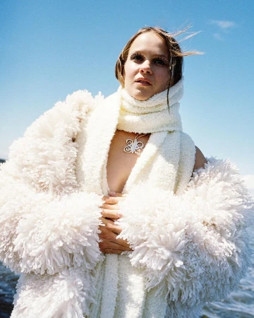 Woman in a fluffy white robe and scarf stands against a blue sky with water in the background, posing with hands together.