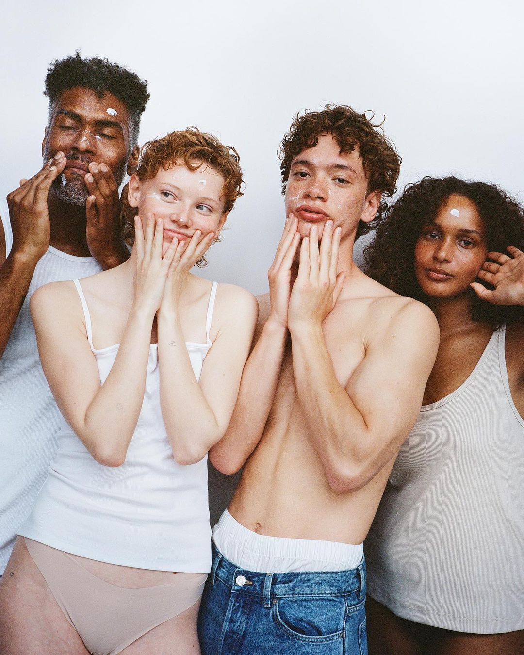 Four multi-ethnic young adults with skincare product on their faces pose in a close-up shot against a white background, smiling gently.