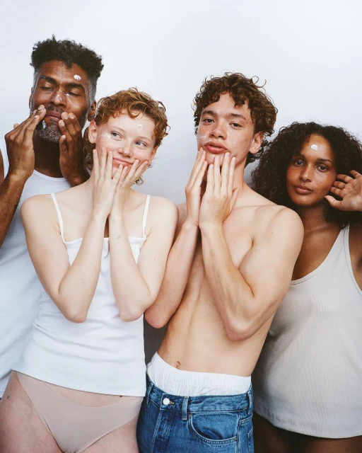 Four multi-ethnic young adults with skincare product on their faces pose in a close-up shot against a white background, smiling gently.