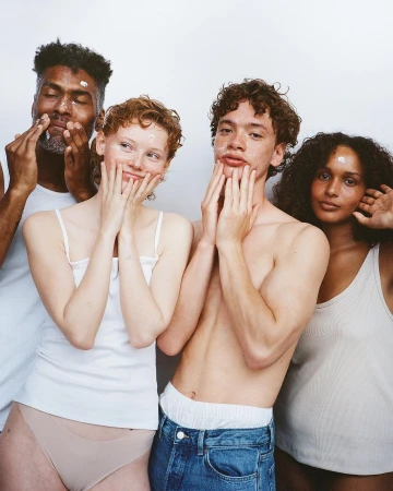 Four multi-ethnic young adults with skincare product on their faces pose in a close-up shot against a white background, smiling gently.