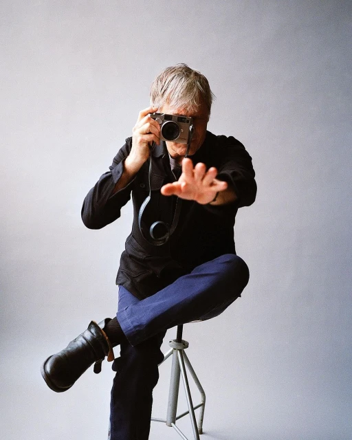 Mature man taking photograph with camera while sitting on metal stool against grey background inside a photo studio.