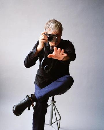 Mature man taking photograph with camera while sitting on metal stool against grey background inside a photo studio.