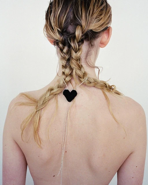 Back view of a fair-skinned adult female with blonde braids, accessorized with a black heart pendant and delicate silver chains.