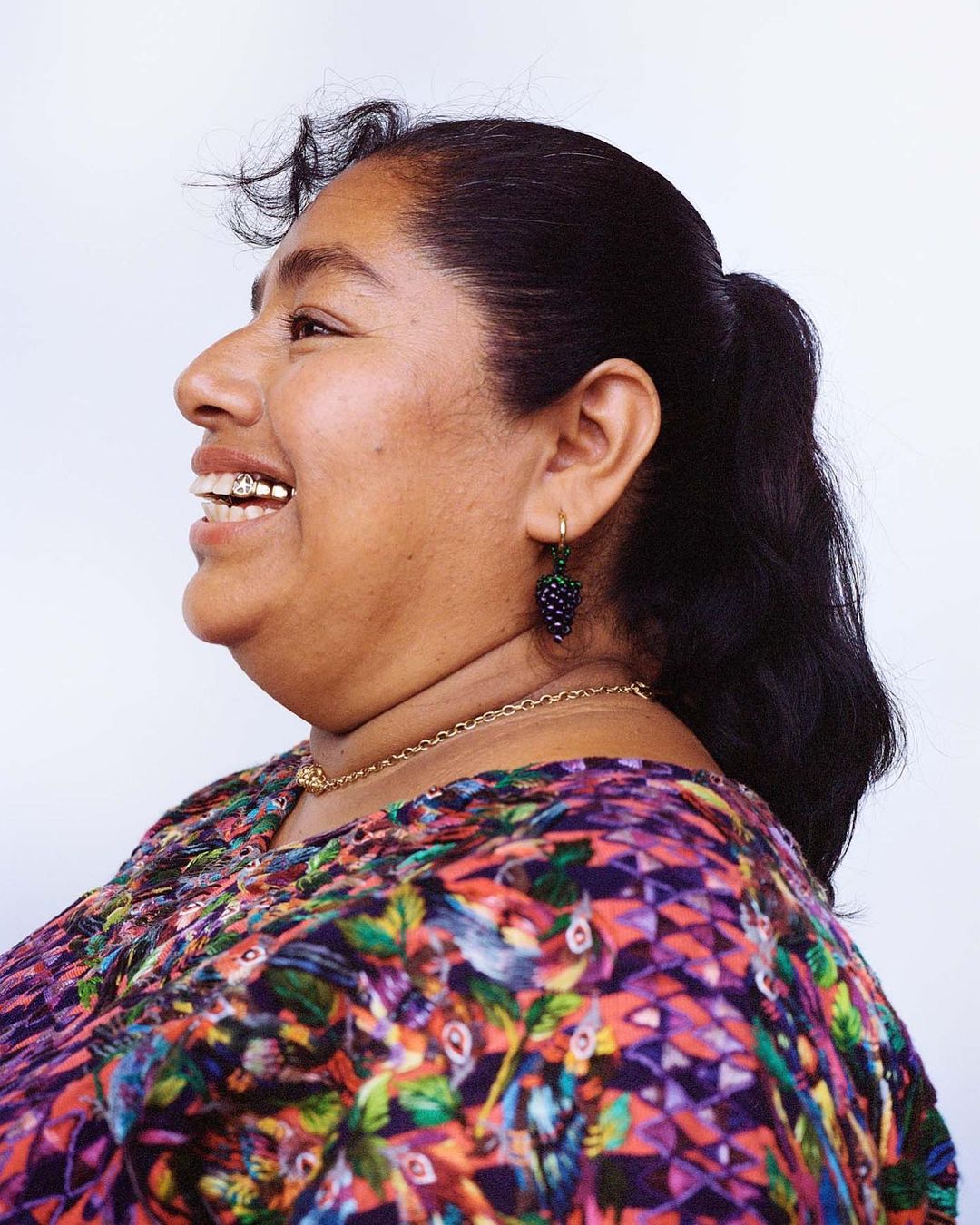 Side view of a smiling adult woman with dark hair in a ponytail, wearing colorful patterned top and grape earrings against a white background.