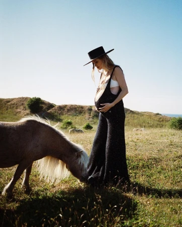 Pregnant woman in black overalls and hat stands in a grassy field, looking at a light brown horse in a rural landscape under a blue sky.