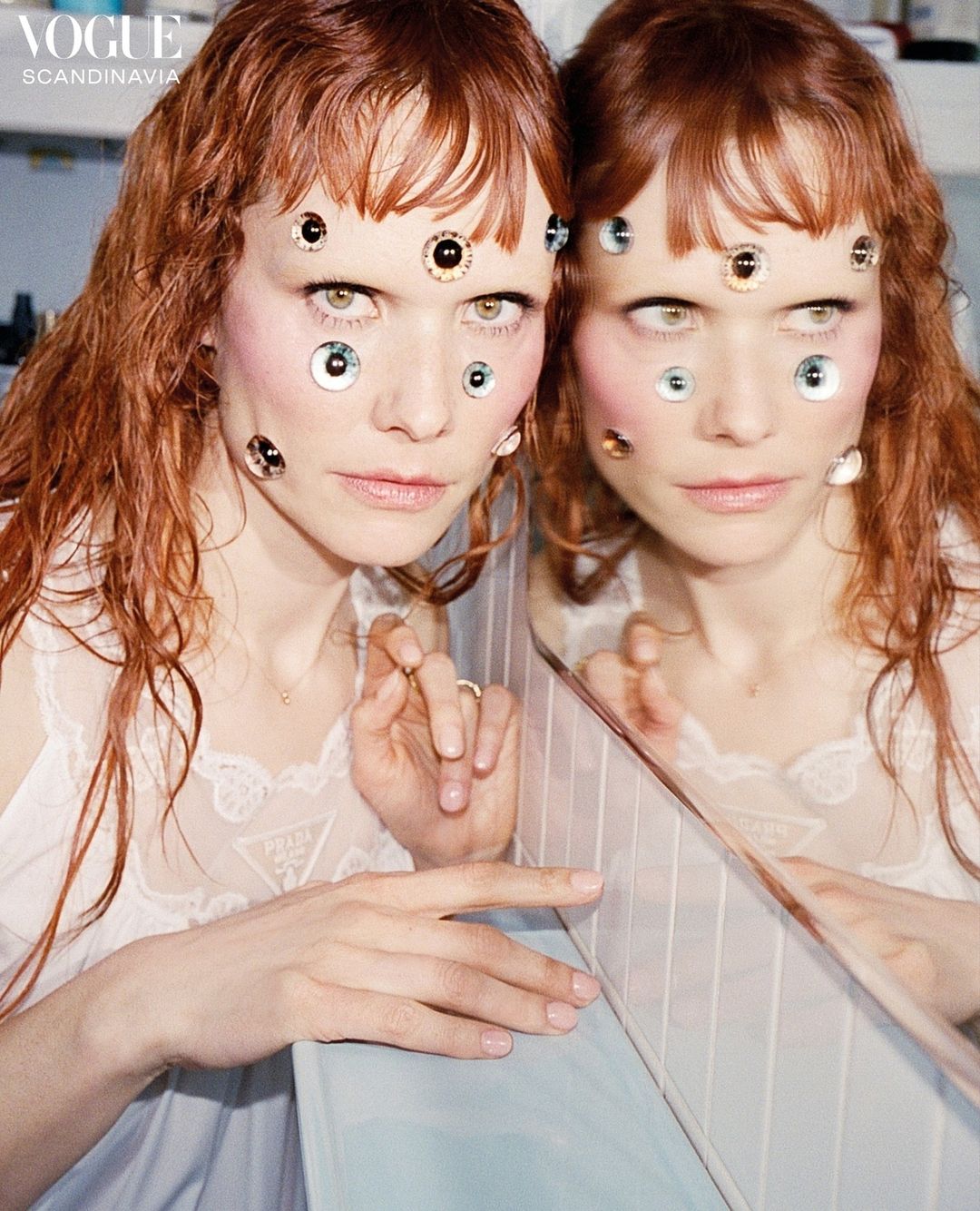 Conceptual fashion portrait: A fair-skinned adult female poses indoors with multiple googly eyes on her face, next to her own reflection.
