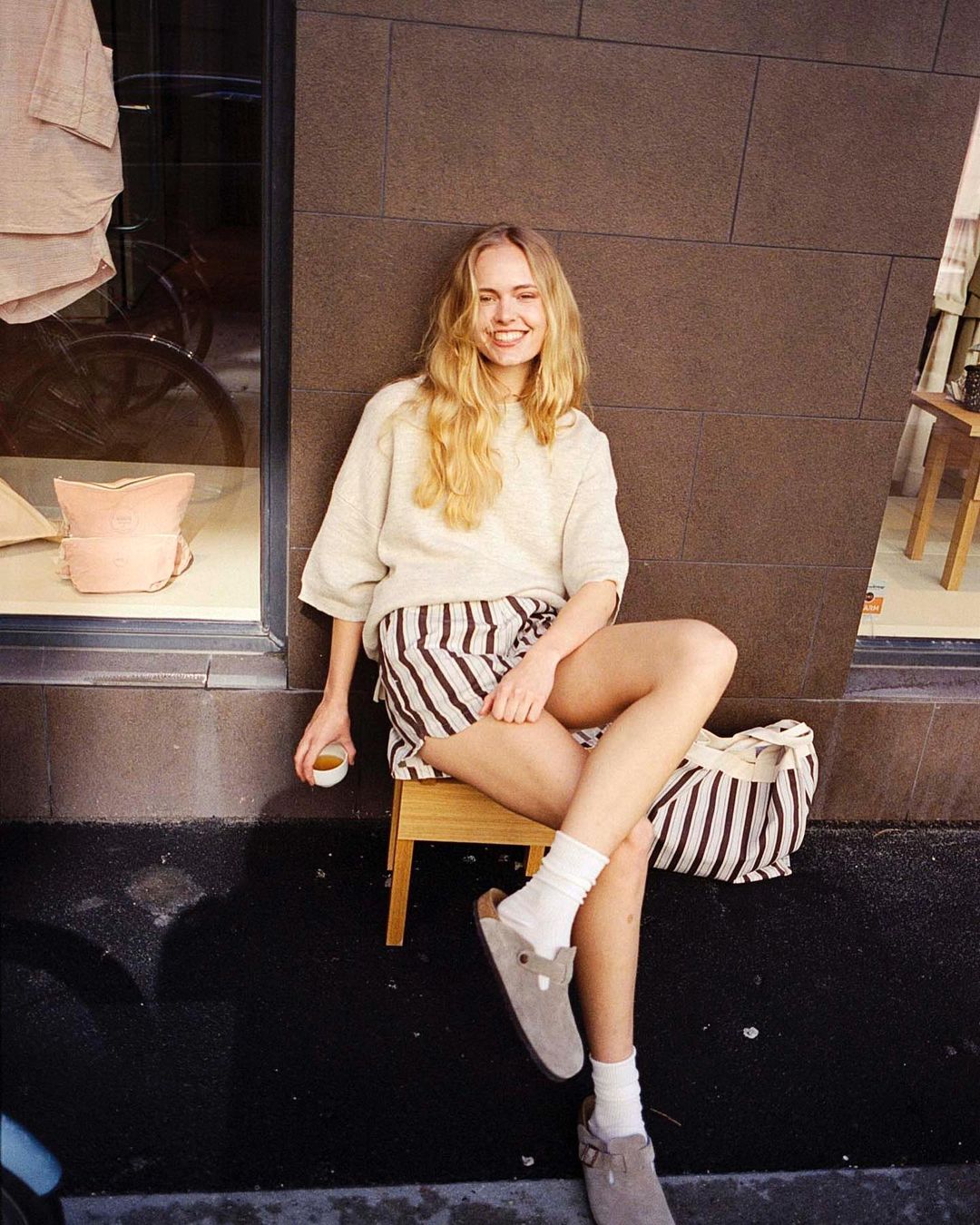 Woman sitting on a stool outdoors in sunlight, wearing striped shorts, a beige sweater, white socks, and clogs, holding a cup. 