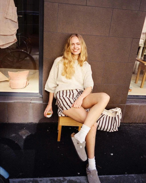 Woman sitting on a stool outdoors in sunlight, wearing striped shorts, a beige sweater, white socks, and clogs, holding a cup. 