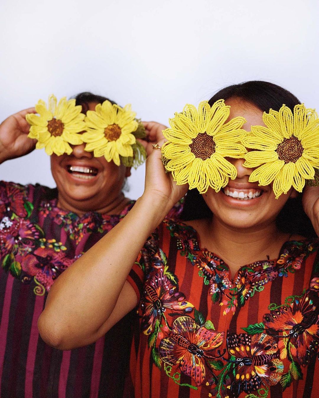 Two smiling women pose indoors with beaded sunflower accessories over their eyes. They wear colorful blouses with striped and floral designs.