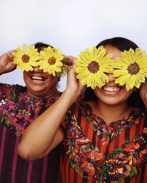 Two smiling women pose indoors with beaded sunflower accessories over their eyes. They wear colorful blouses with striped and floral designs.