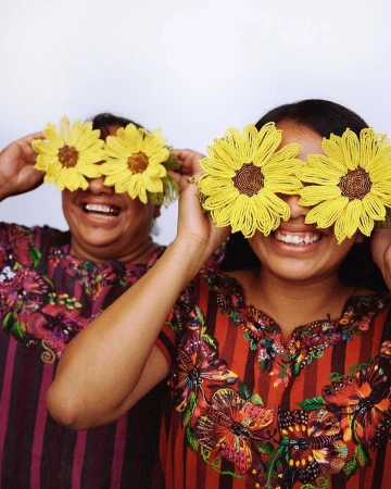 Two smiling women pose indoors with beaded sunflower accessories over their eyes. They wear colorful blouses with striped and floral designs.