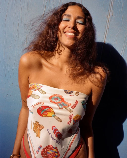 Smiling adult woman with brown curly hair wearing a patterned scarf around her chest, with a blue wall in the background.