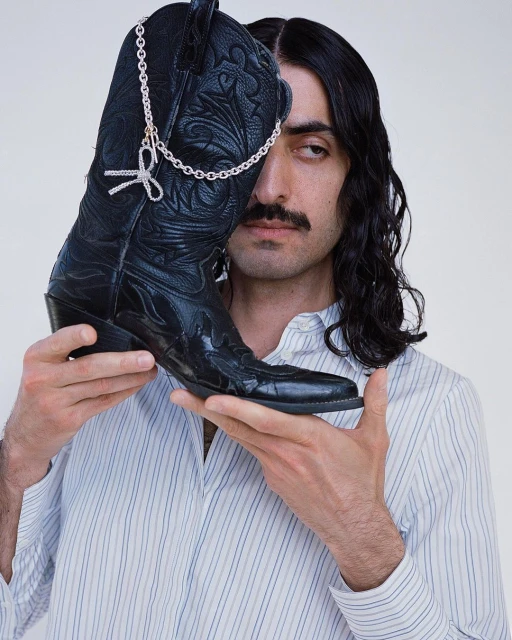 Adult male with mustache wearing a button-down shirt, holding a black cowboy boot decorated with a silver chain and bow.