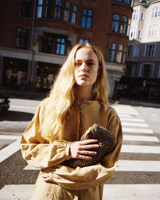 A young woman in a yellow dress poses on a city street near a crosswalk, holding a brown textured purse in a medium shot.