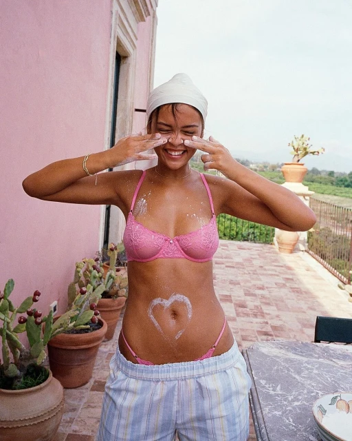 Smiling woman with pink bra and boxer shorts poses outside with sunscreen on her face and stomach, near cacti and greenery.