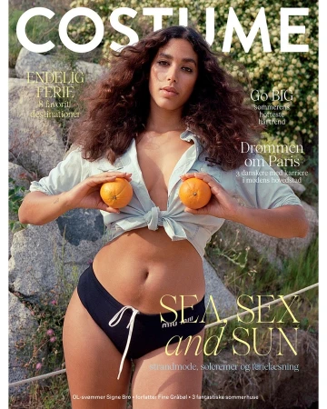 Woman with long curly brown hair holding two oranges against her chest, wearing a tied light blue shirt and dark briefs outdoors.
