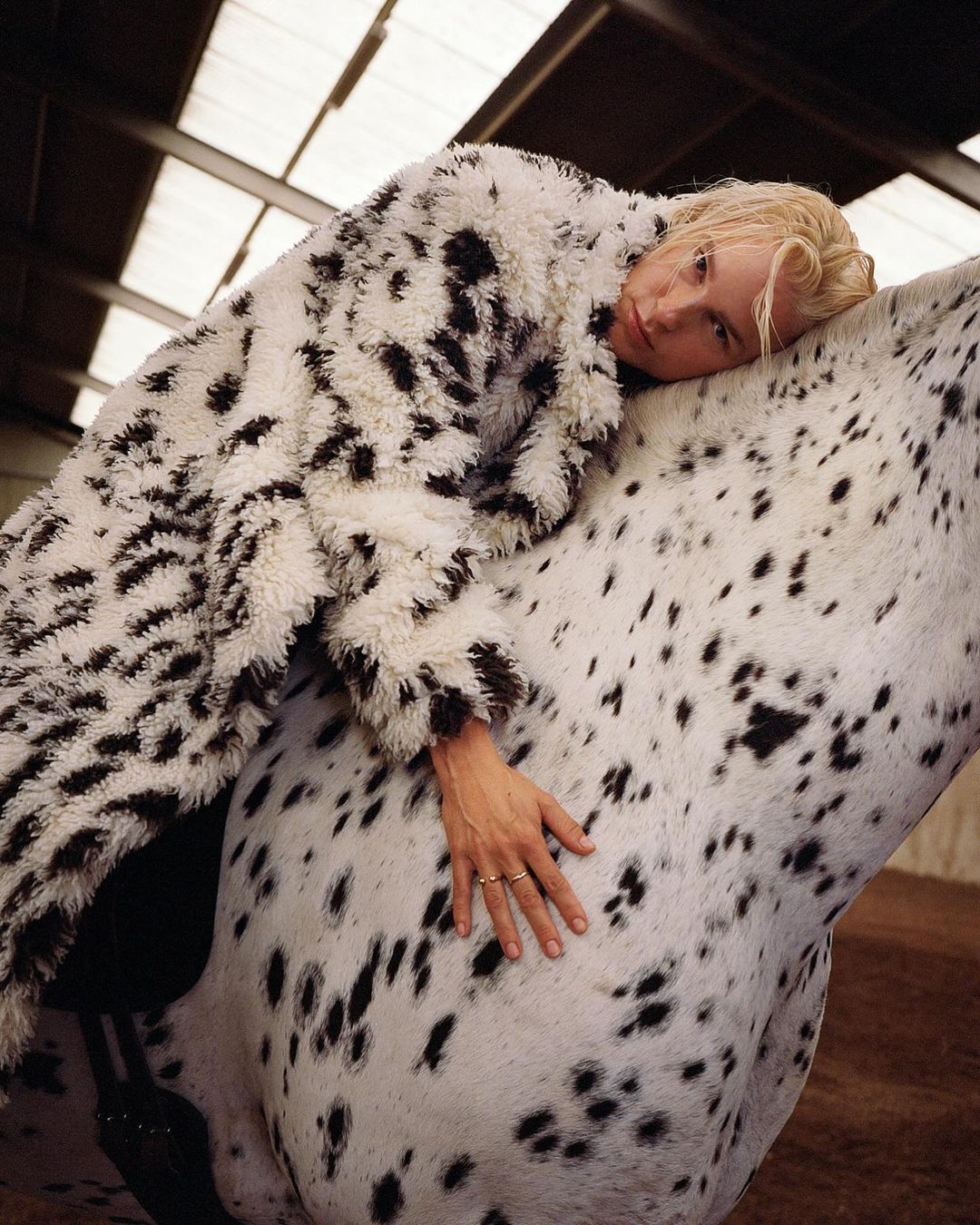 Woman in a spotted coat lies on a white horse with black spots in an indoor setting with light coming from the ceiling.