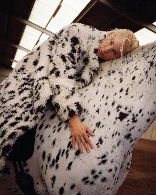 Woman in a spotted coat lies on a white horse with black spots in an indoor setting with light coming from the ceiling.