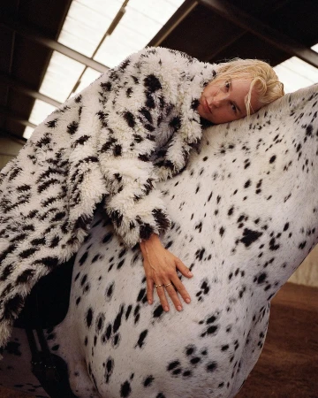 Woman in a spotted coat lies on a white horse with black spots in an indoor setting with light coming from the ceiling.