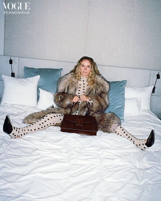 A woman in a fur coat and polka dot tights sits on a bed, holding a brown bag, in a fashion shot for Vogue Scandinavia.