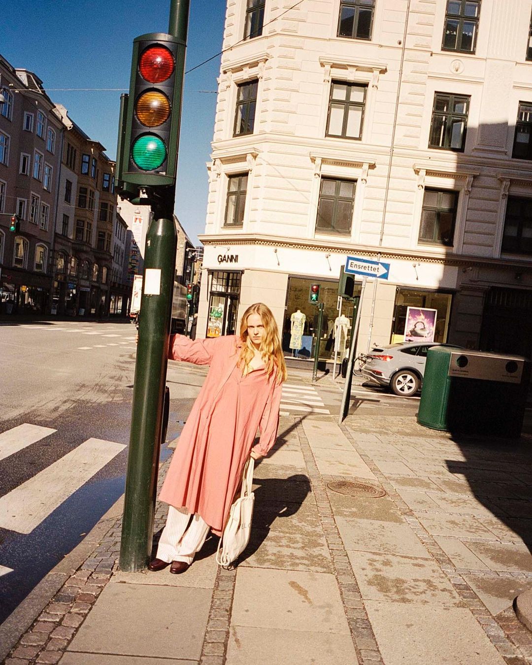 A fashionable adult female stands on a city sidewalk next to a traffic light, wearing a pink dress and carrying a tote bag in daylight.