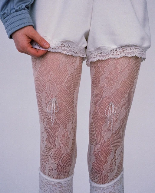 A woman is posing with white lace tights, wearing white shorts with a lace trim and a light-blue top in front of a grey background.