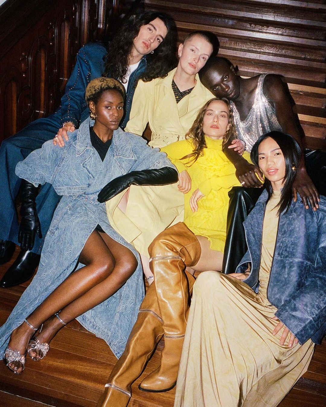 A group of six fashion models pose indoors wearing denim and tan outfits; light skin tones and dark skin tones are featured in the group.