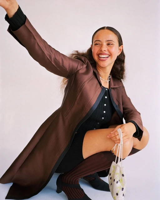 Young woman in a brown coat smiles while crouching in a studio setting, holding a beaded purse, exuding a happy and stylish vibe.