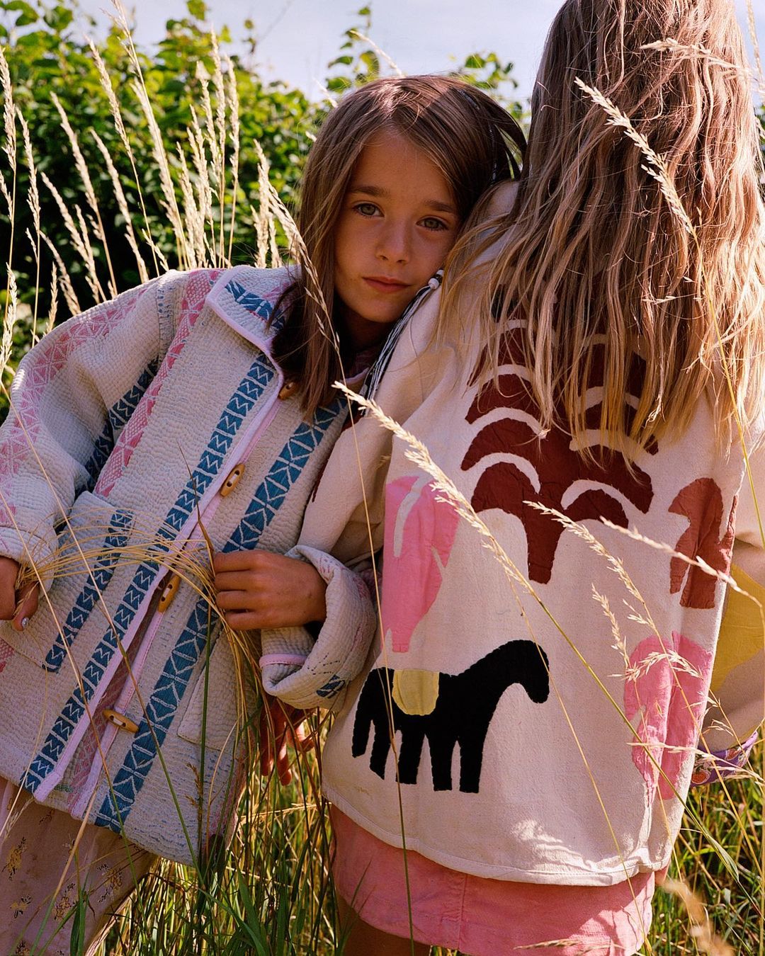 Two young girls pose outdoors in embroidered jackets, surrounded by tall grass and foliage in a natural setting.