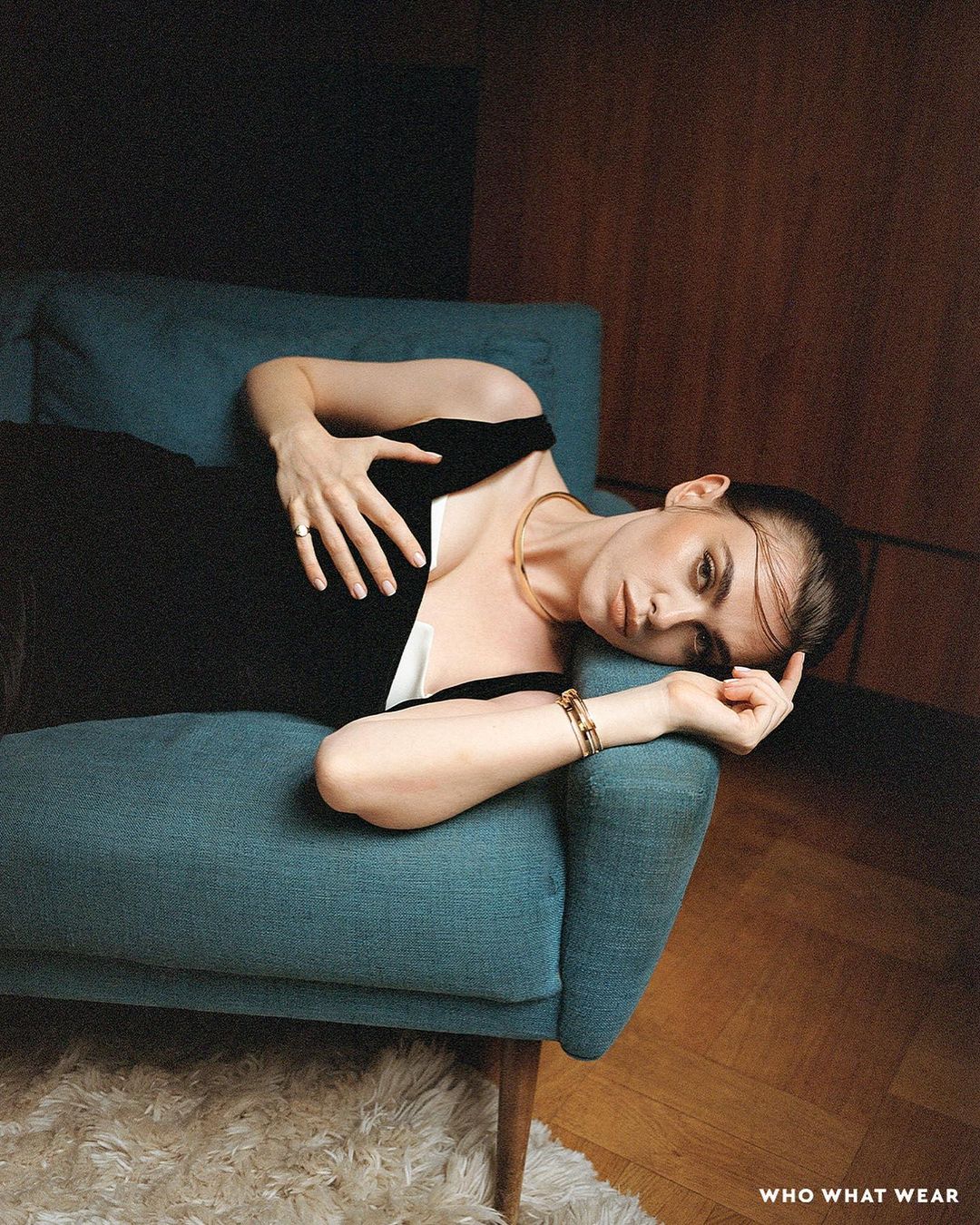 A woman wearing a black dress and gold necklace reclines on a teal couch indoors with a wood floor in a fashion editorial style.