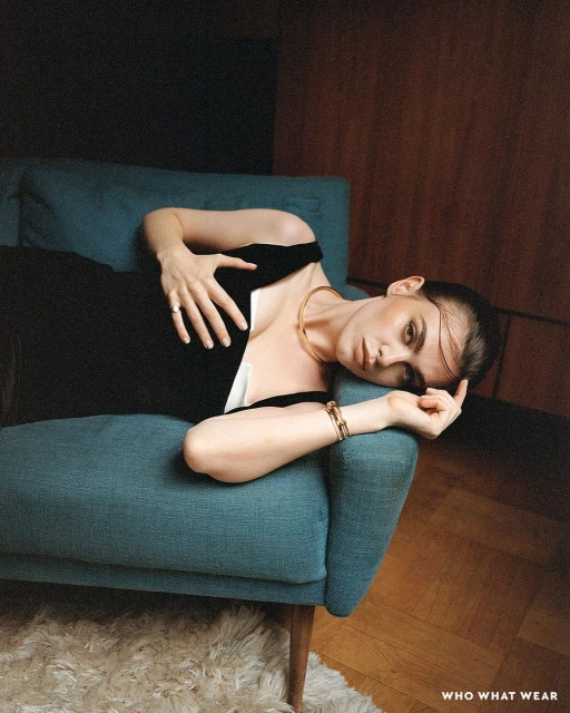 A woman wearing a black dress and gold necklace reclines on a teal couch indoors with a wood floor in a fashion editorial style.