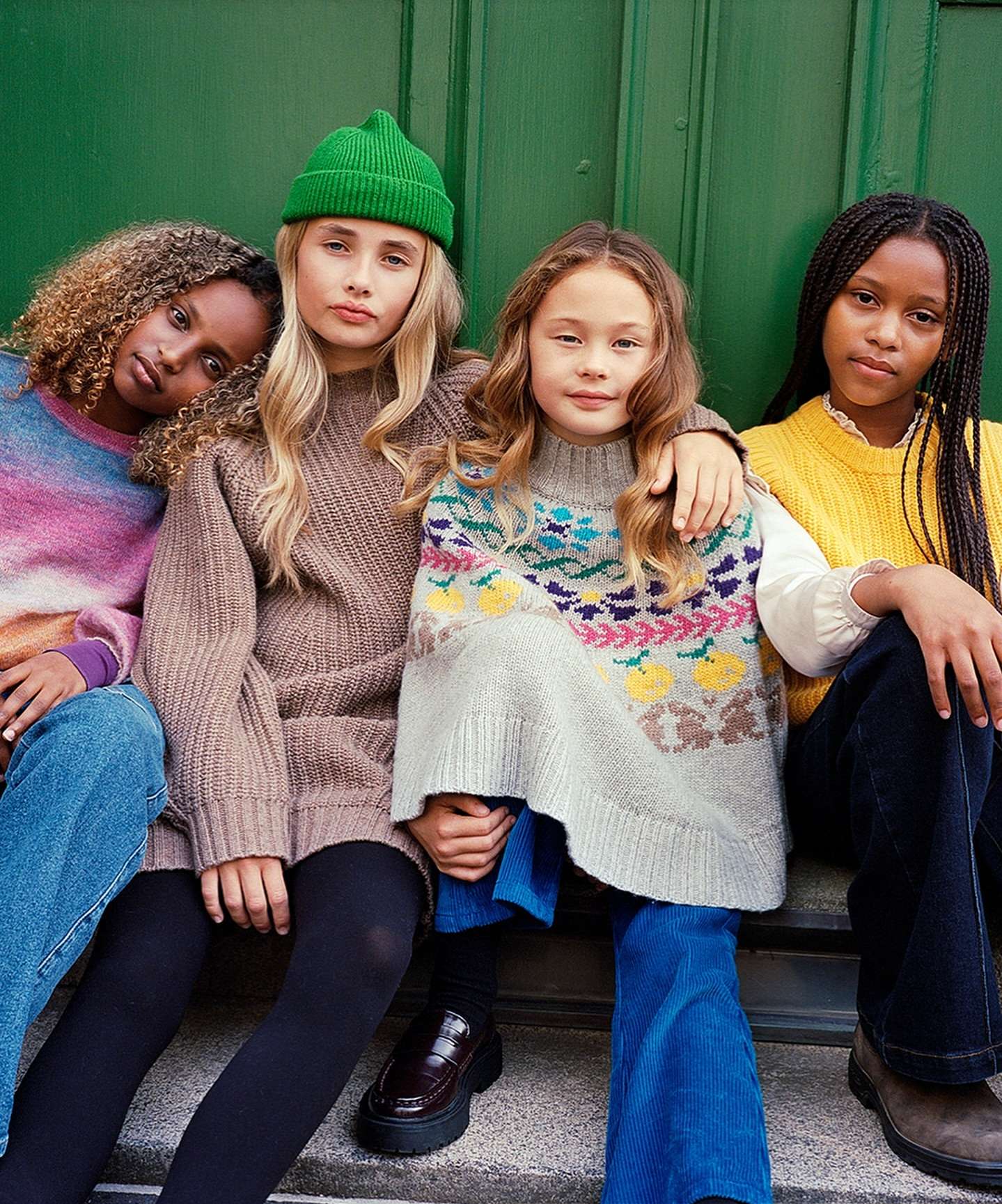 Four young girls in fashionable knitwear and denim pose against a green door, showcasing a blend of textures and stylish designs.