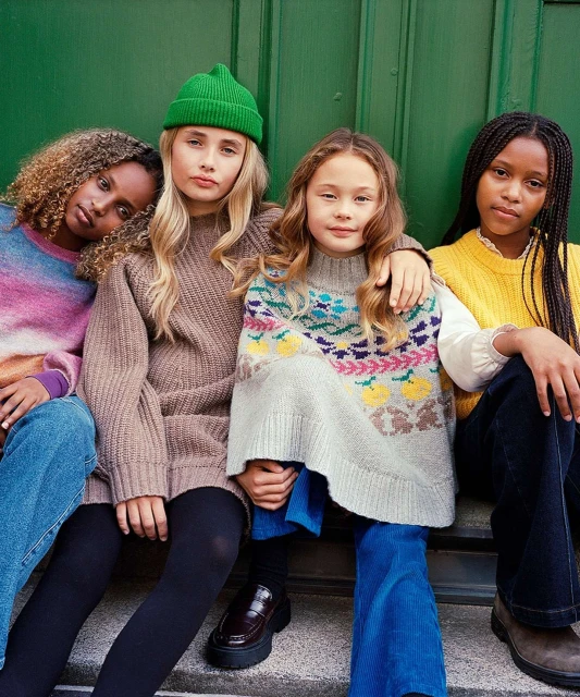 Four young girls in fashionable knitwear and denim pose against a green door, showcasing a blend of textures and stylish designs.