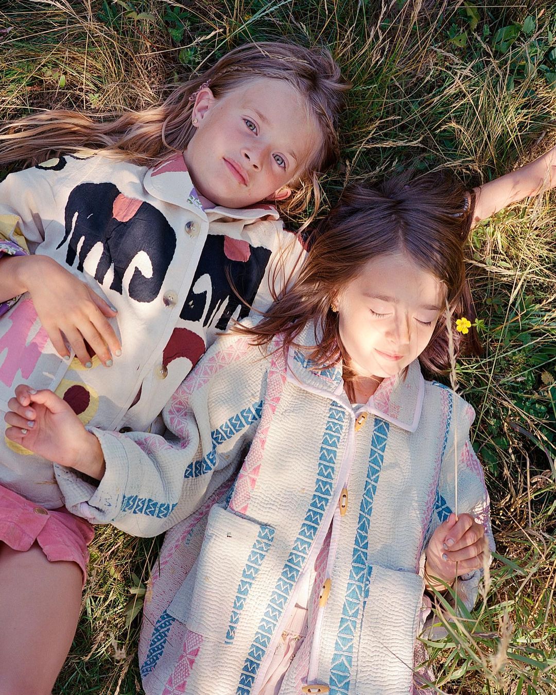Two young girls in patterned jackets lie in a grassy field with a single yellow flower, enjoying the sunlight in a close-up shot.