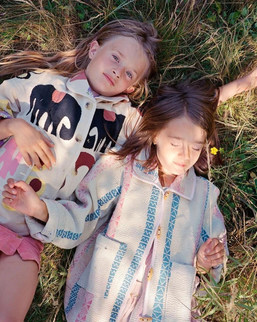 Two young girls in patterned jackets lie in a grassy field with a single yellow flower, enjoying the sunlight in a close-up shot.