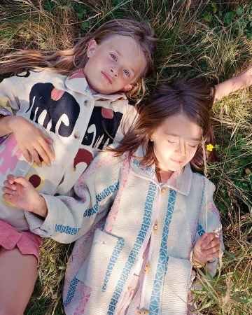 Two young girls in patterned jackets lie in a grassy field with a single yellow flower, enjoying the sunlight in a close-up shot.