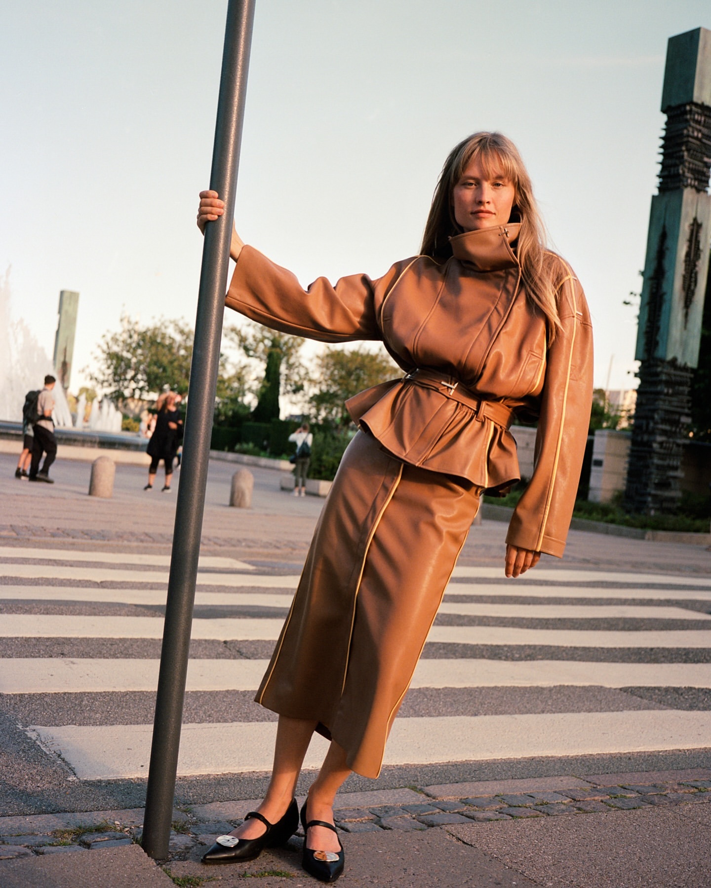 Fashionable woman poses in a brown leather skirt suit while leaning against a pole on a crosswalk in an urban environment. 