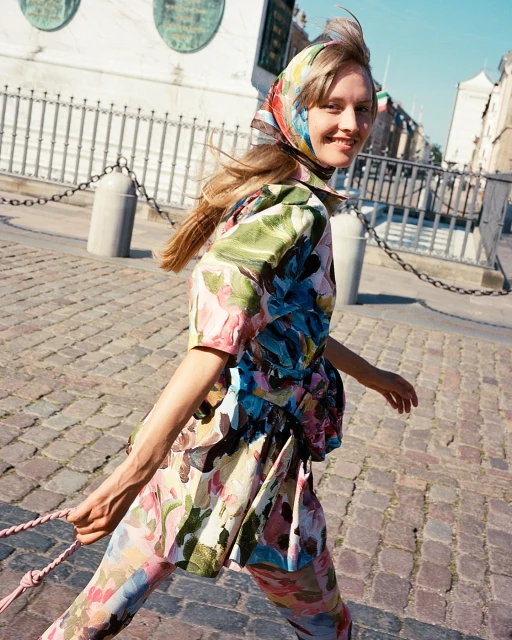 Adult woman in floral print outfit and headscarf walks on a cobblestone street near a building in an outdoor urban setting.