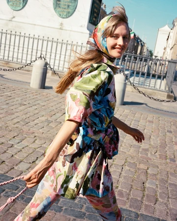 Adult woman in floral print outfit and headscarf walks on a cobblestone street near a building in an outdoor urban setting.